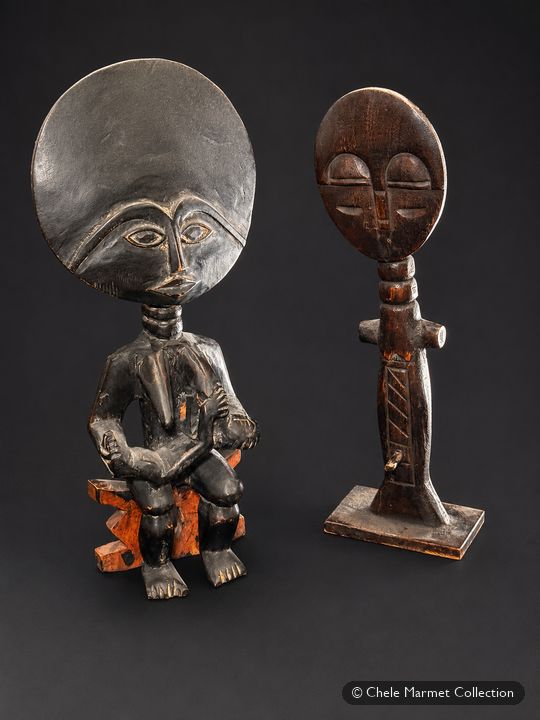 Two dark carved figures with large, round, flat heads: one seated and holding a baby at the breast, one standing.