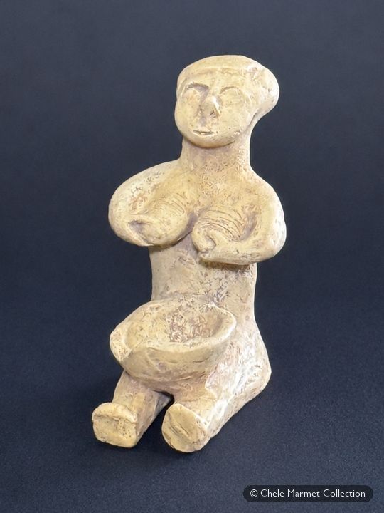 Small pale seated figure pressing both hands to her breasts.