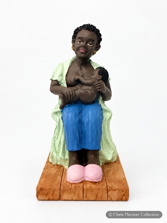 Painted figure of a Black mother in a green blouse, blue trousers and pink slippers, seated on a wooden base and nursing a baby.