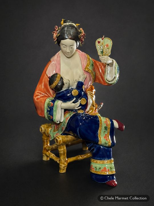 Glazed ceramic figure of a woman in an orange and blue robe, seated on a bamboo stool and nursing a baby.