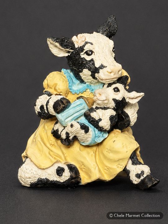 Painted figurine of a cow in a yellow skirt giving a bottle to a calf.