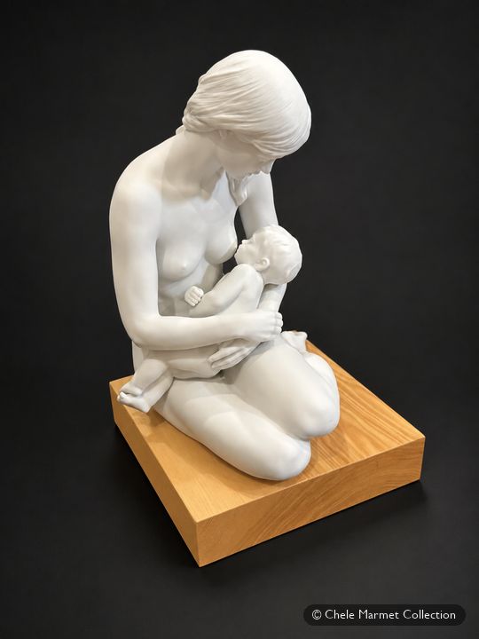 White porcelain figure of a kneeling woman nursing a baby, on a wooden base.