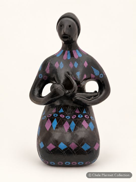 Black glazed ceramic figure of a woman with a long neck and small face, decorated with pink and blue diamonds, holding a baby to her breast.