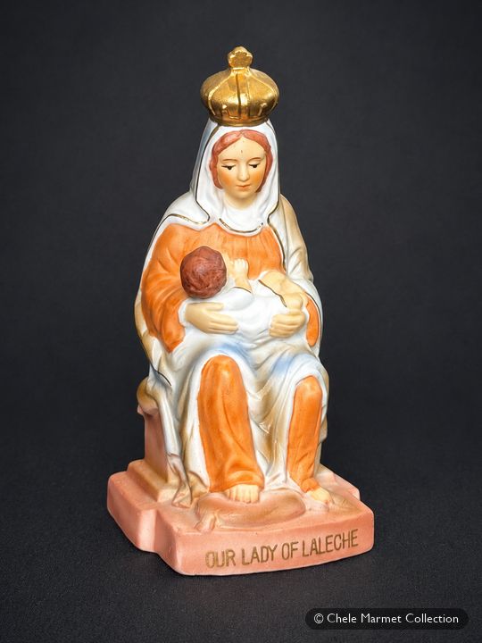 Painted statuette of a crowned Madonna in a white and orange robe nursing a child, on a pink base lettered “Our Lady of La Leche.”