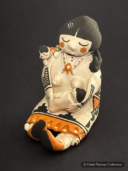 Small white painted pottery figure with black hair and a patterned skirt, nursing a baby.
