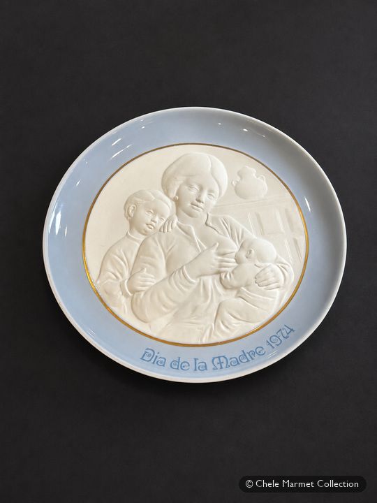 Pale blue plate with a white relief of a mother nursing a baby while a child leans on her shoulder, lettered “Día de la Madre 1974.”