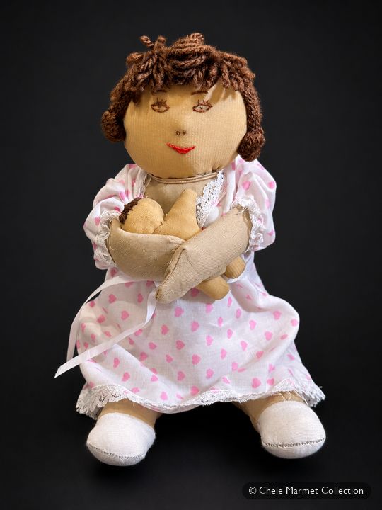 Cloth doll with brown yarn hair in a pink dotted dress, holding a cloth baby at her breast.